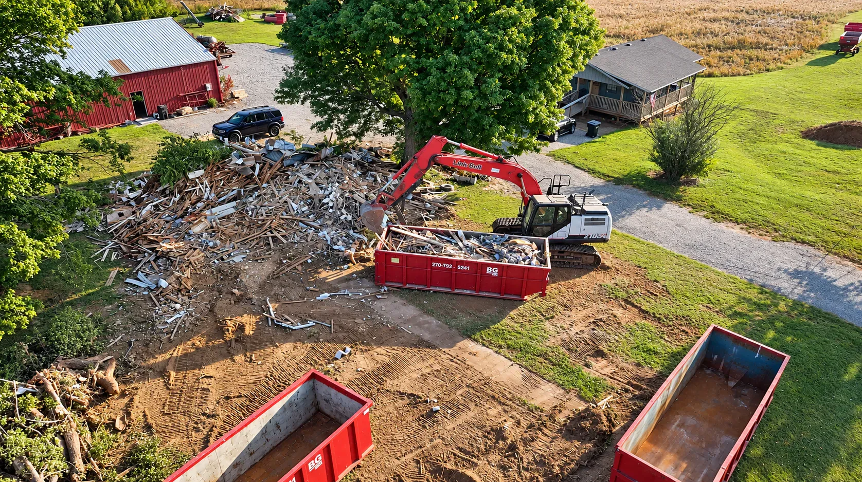 An excavator loading demolition debris into a red BG Dumpster roll-off container at a rural property